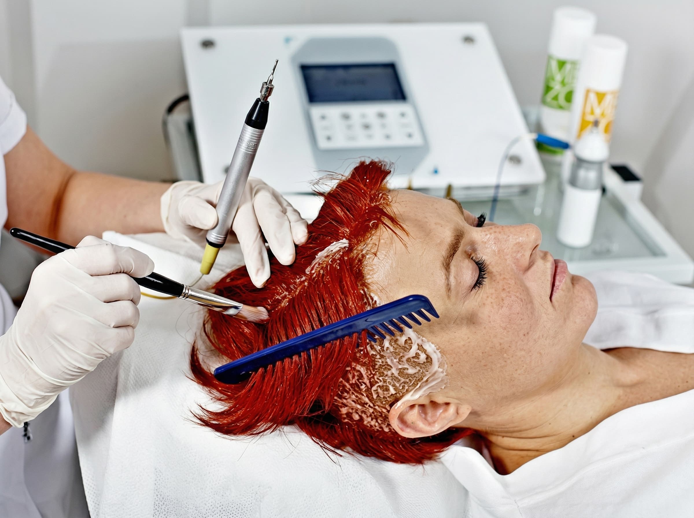 Trycholog w&nbsp;białych rękawiczkach aplikuje specjalistyczny preparat na&nbsp;skórę głowy pacjentki używając pędzelka. W&nbsp;tle widoczne nowoczesne urządzenie do&nbsp;zabiegów i&nbsp;profesjonalne kosmetyki w&nbsp;gabinecie Rokoko.