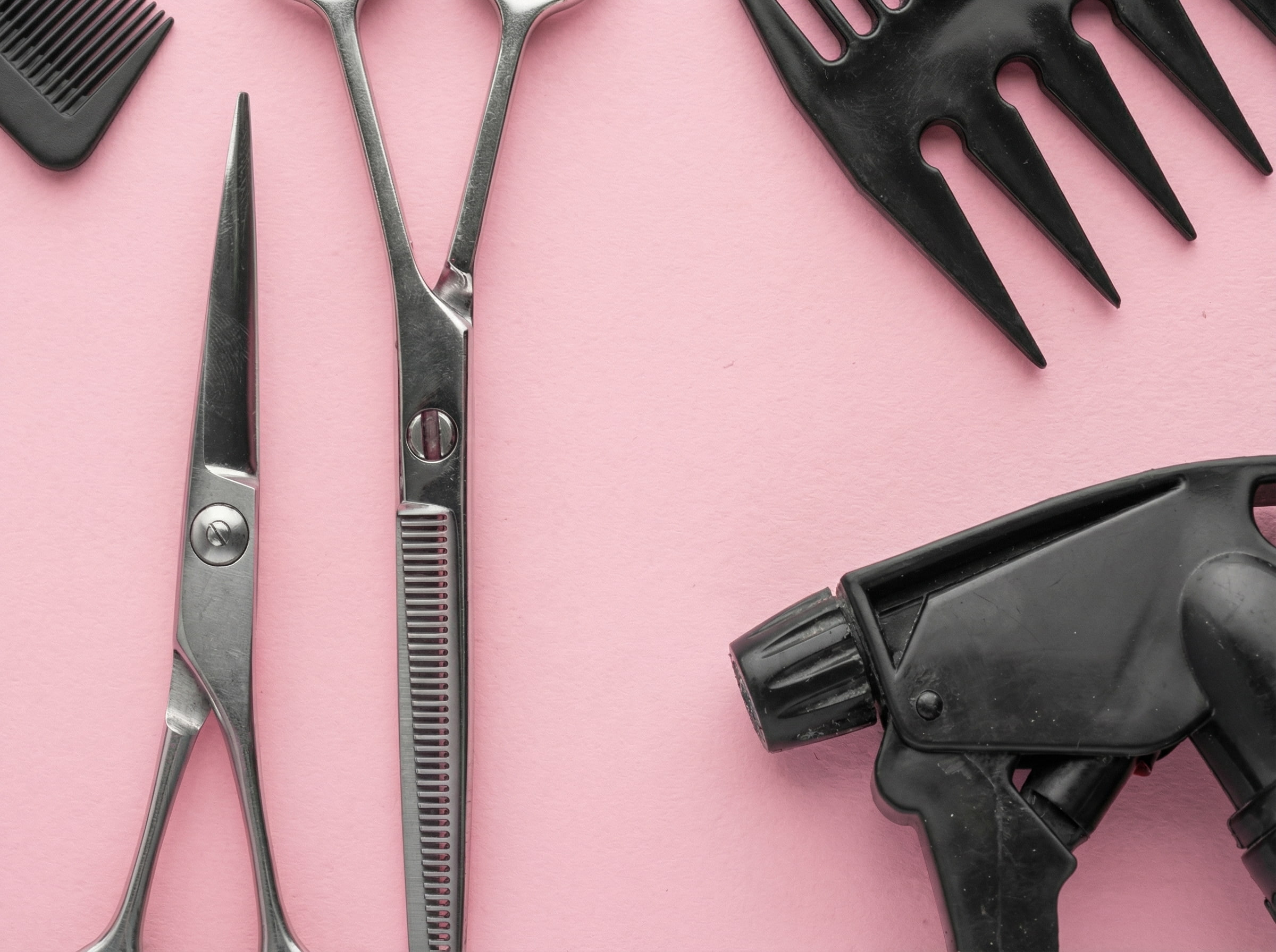 Zestaw czarnych i&nbsp;metalowych narzędzi fryzjerskich: nożyczki, spryskiwacz, klipsy i&nbsp;grzebienie, estetycznie ułożonych na&nbsp;pastelowym, różowym tle.