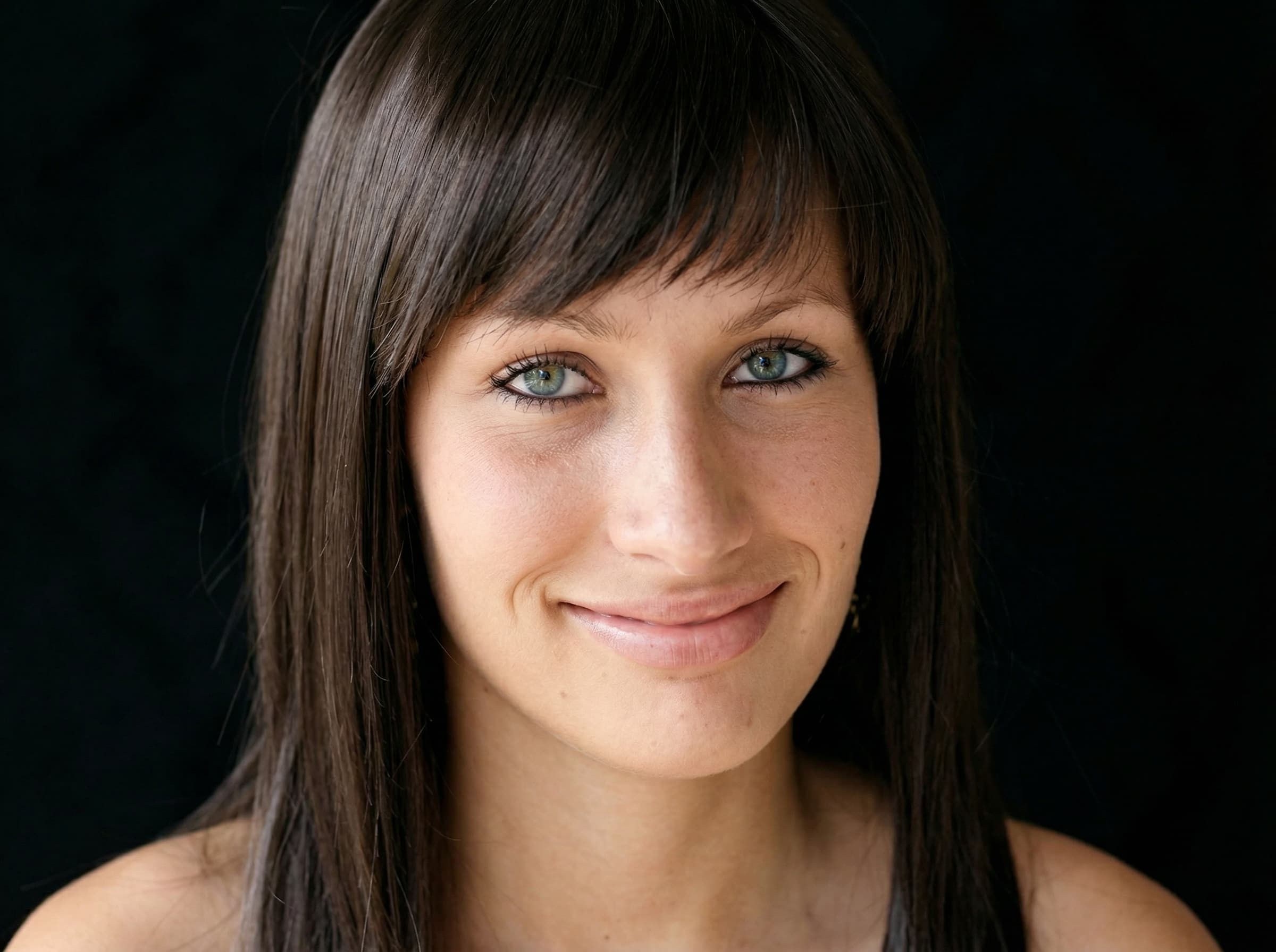 Portret uśmiechniętej kobiety z&nbsp;niebieskimi oczami w&nbsp;peruce z&nbsp;ciemnobrązowych, prostych włosów z&nbsp;grzywką na&nbsp;czarnym tle.