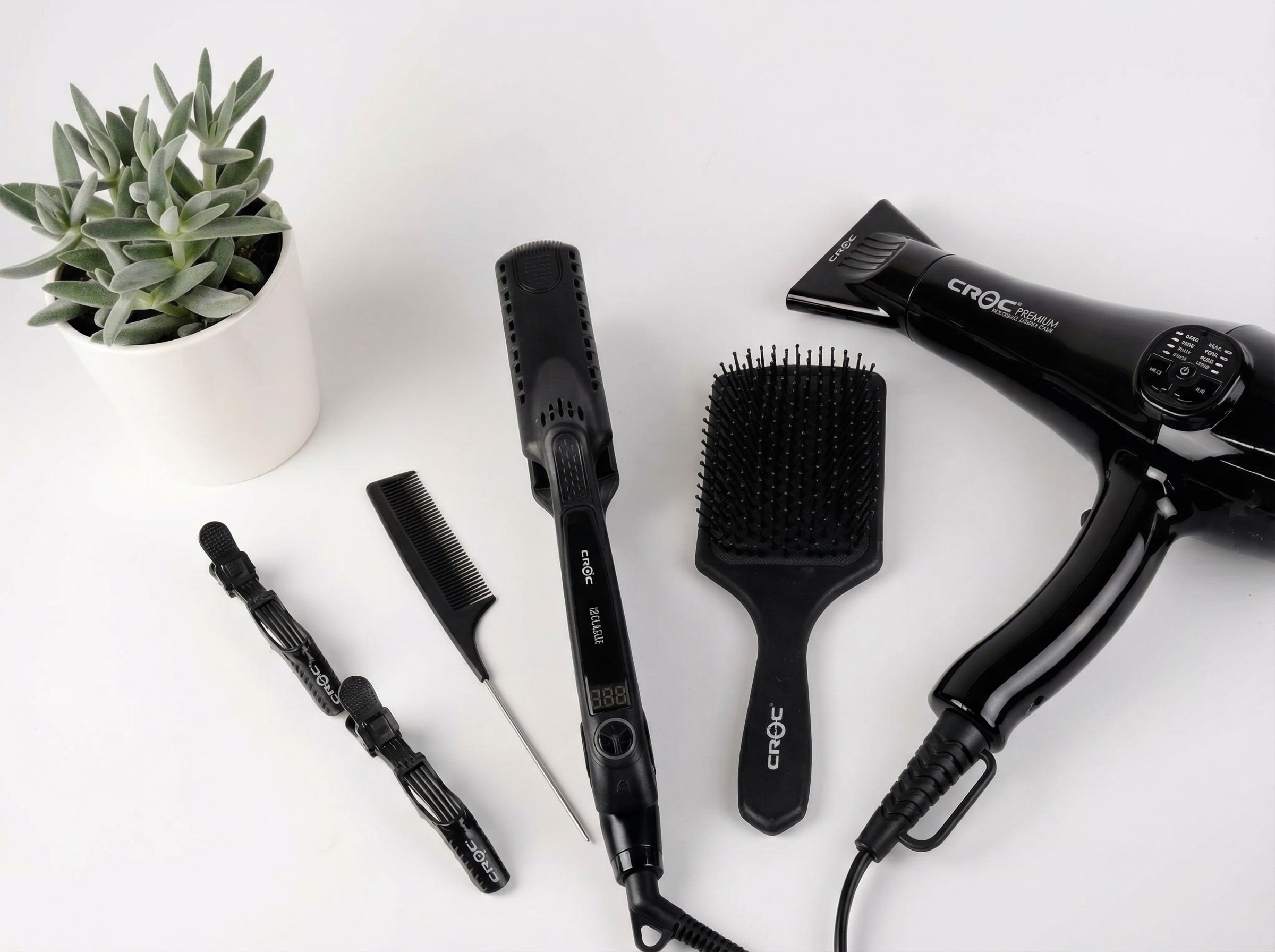Czarna suszarka, karbowana lokówka, szczotka i&nbsp;grzebień ułożone na&nbsp;białym stole obok małej, zielonej rośliny w&nbsp;doniczce.